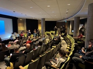 Auditorium with green chairs and a stage left; people seated and some standing near the back during a presentation to an audience. -or- A lecture hall filled with attendees, a projected screen on the left, and several people standing at the back.