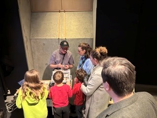 Instructor demonstrates a hands-on activity to children and adults gathered around a table in a workshop setting.