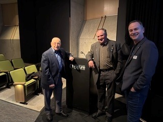 Three men in formal attire stand on a theater stage beside a podium, smiling for the camera.