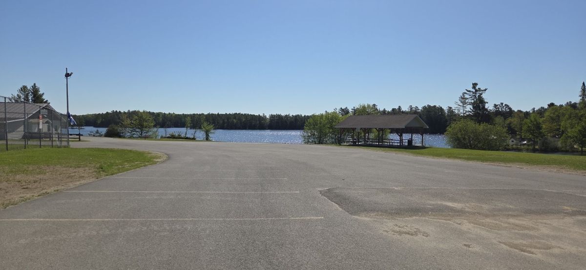 Empty parking lot facing a calm lake with a wooden pavilion and green trees under a clear blue sky